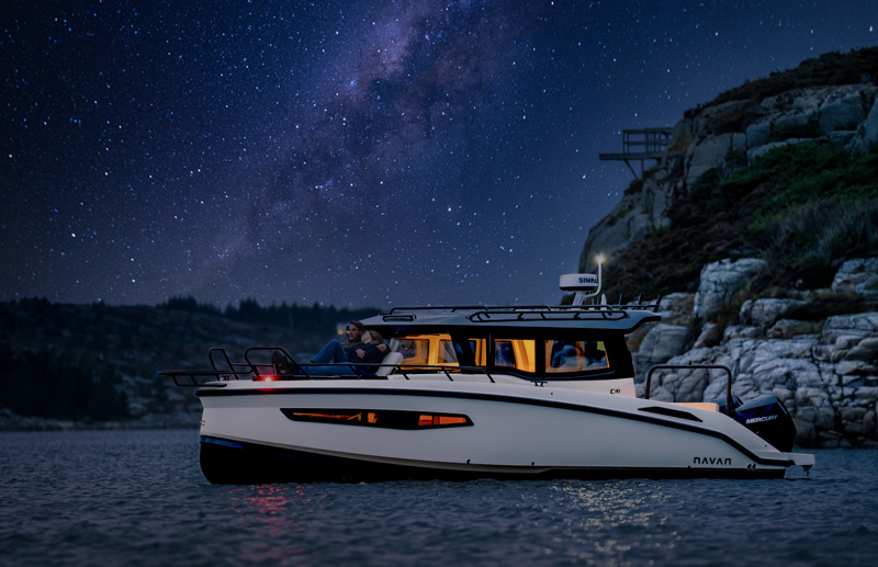 Navan C30 at anchor by a rocky cliff at night, warm cabin light under a starry sky